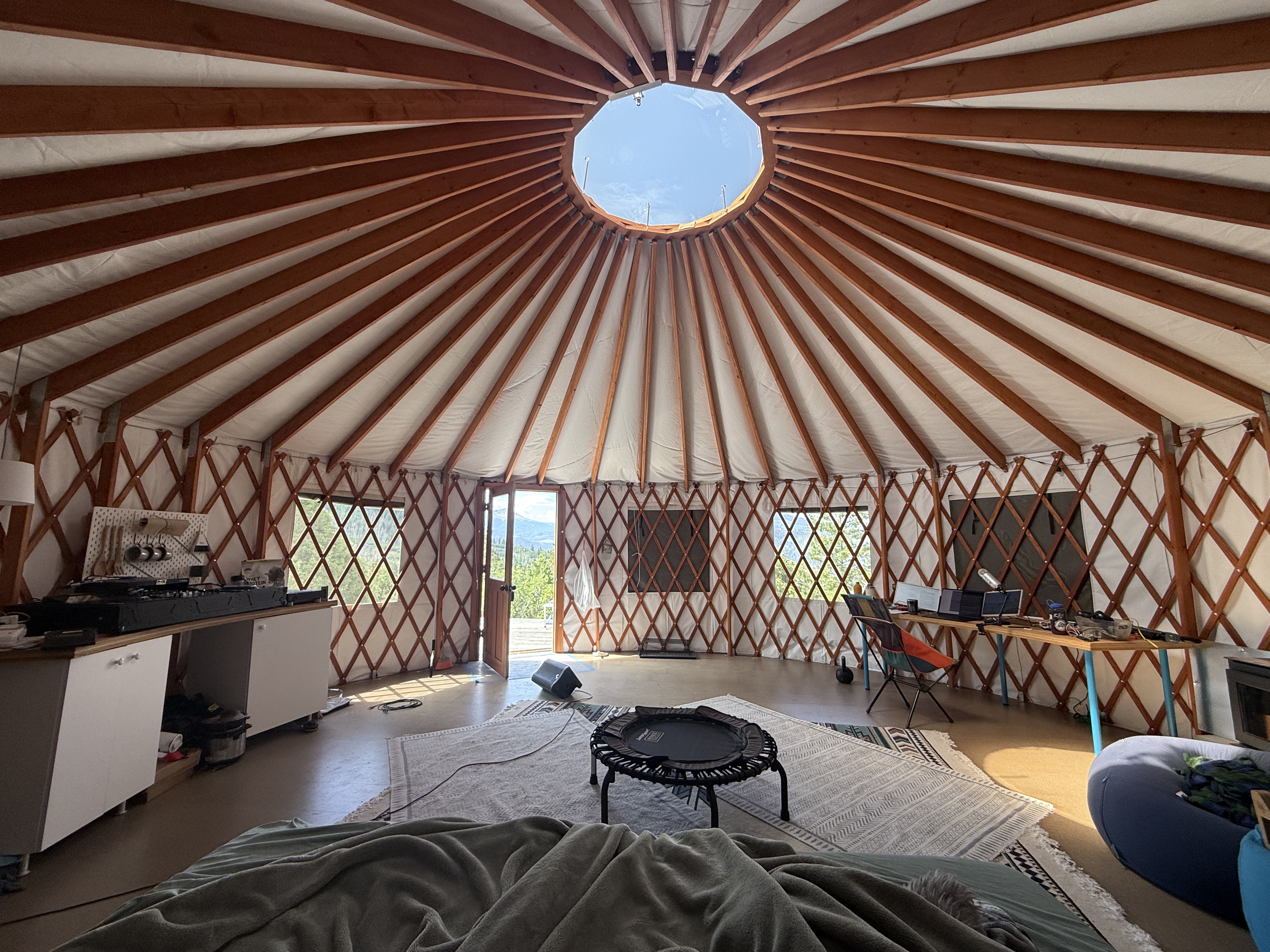 Interior of the yurt above Telluride: lattice walls, dome skylight open to blue sky, music gear along the left wall, desk and equipment on the right, mini trampoline in the center, mountain view through the open door.