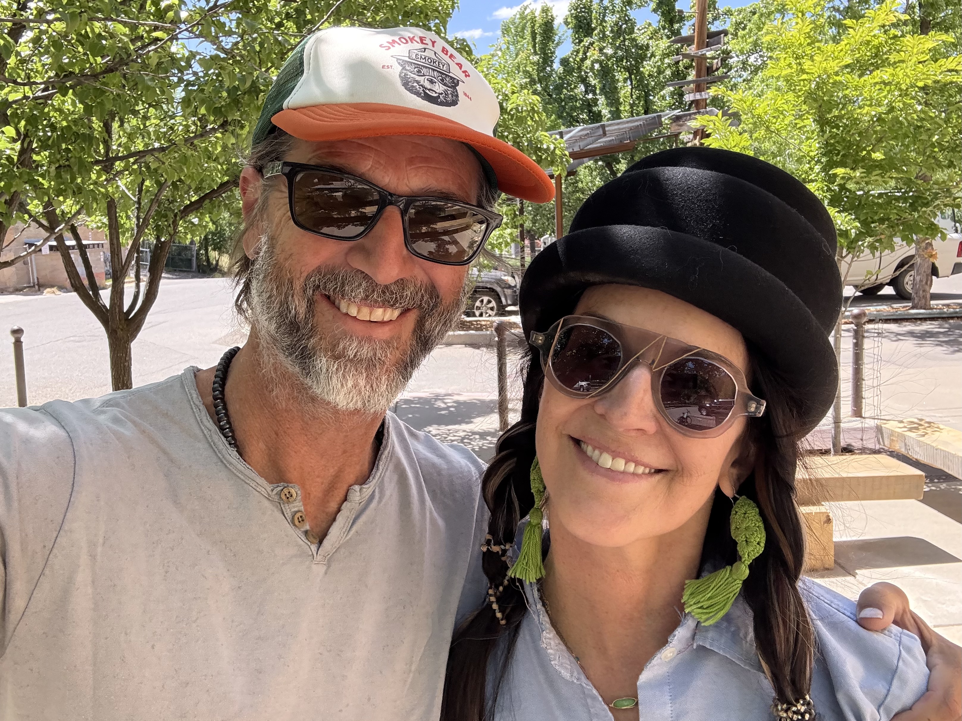Two old friends reuniting in Durango, Colorado after 35 years. Smiling together at an outdoor spot downtown, green trees and blue sky behind them.