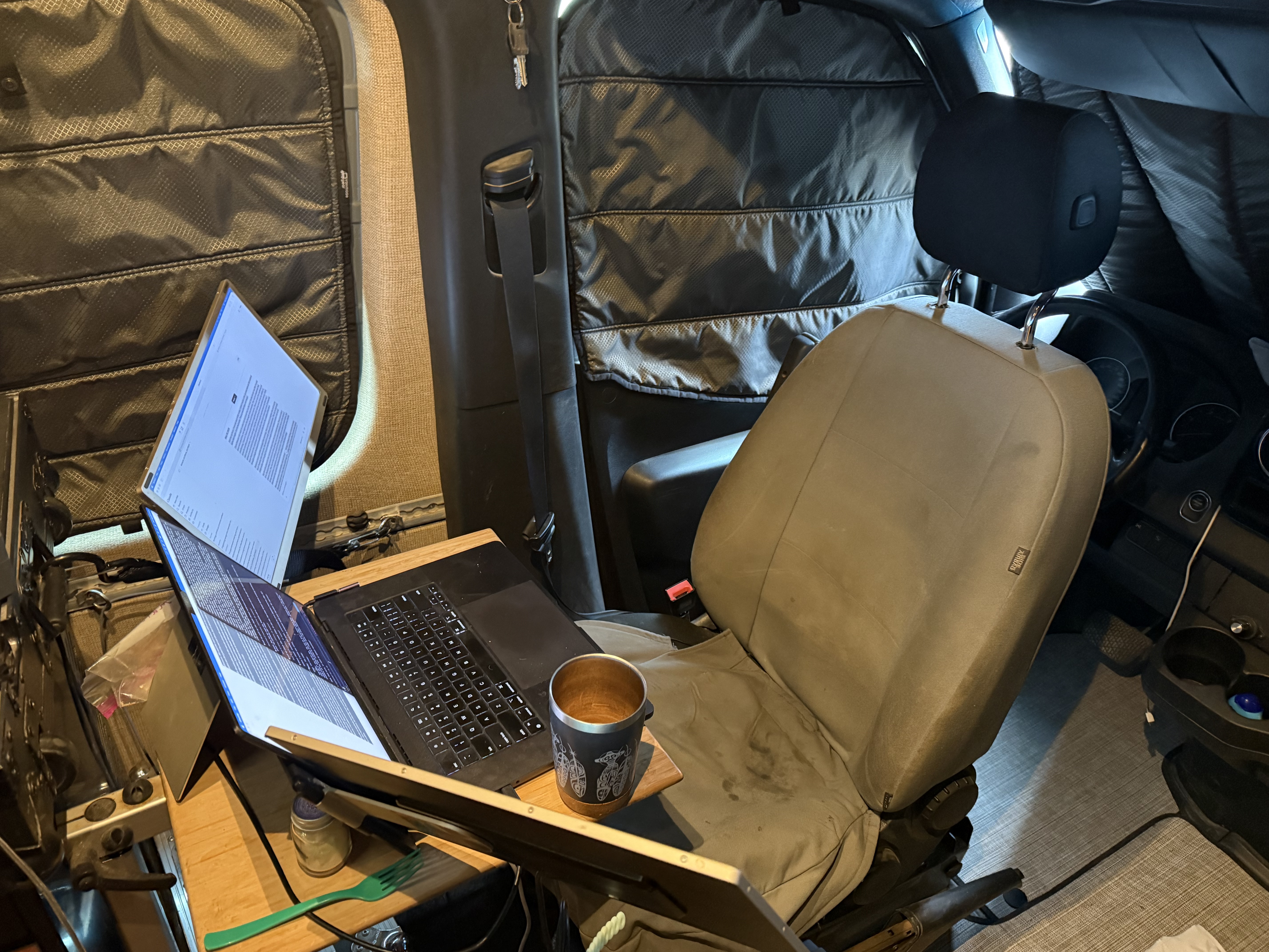 Interior of a Sprinter van set up as a mobile workspace: laptop open on a bamboo tray table beside the passenger seat, steel coffee thermos, green fork