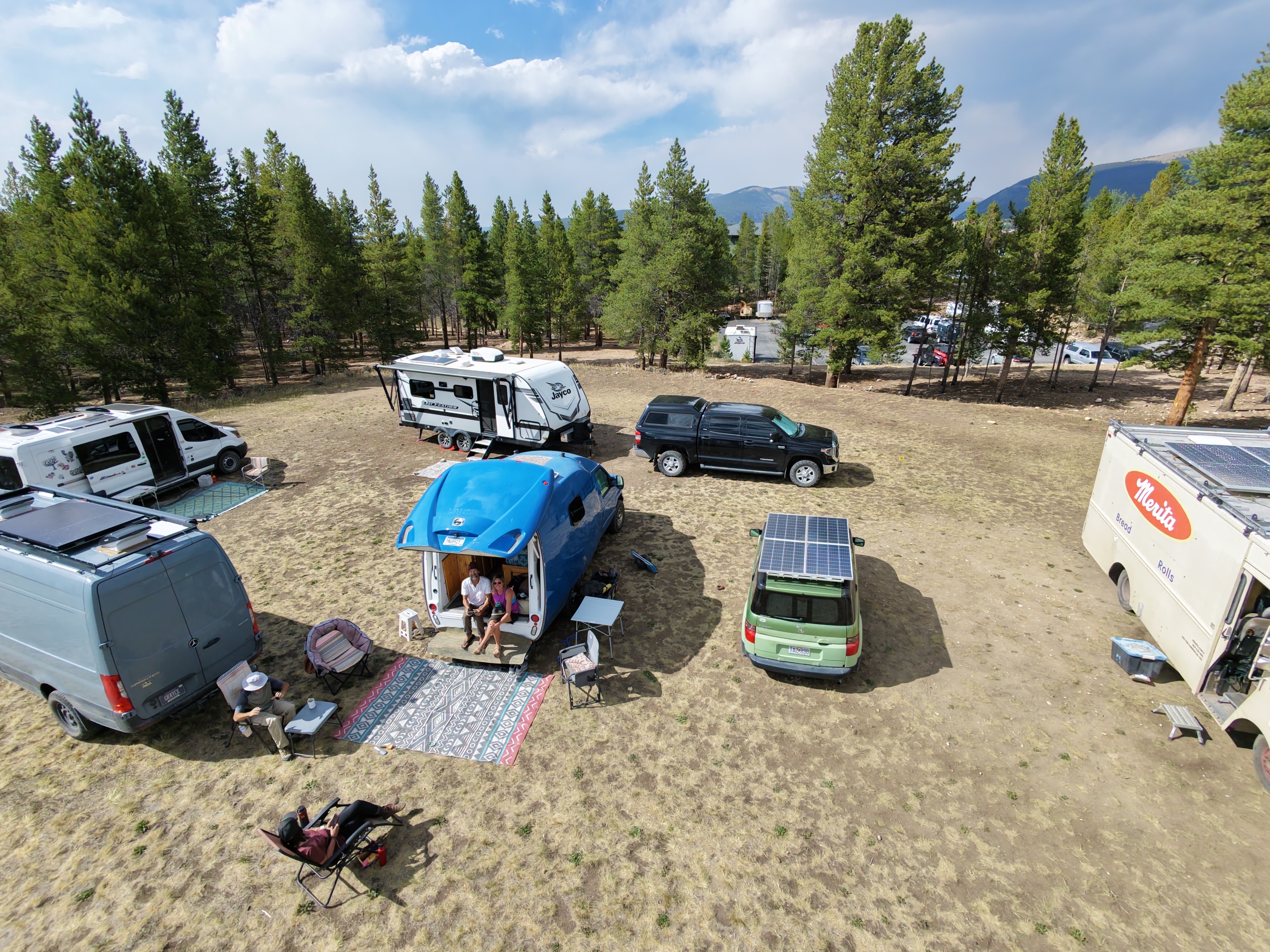 Aerial drone shot of the nomad camp in Leadville, Colorado, showing a cluster of vehicles including a Merita bread truck, green Honda Element, and blue vans in a pine forest clearing, with wildfire smoke rising from distant mountains.