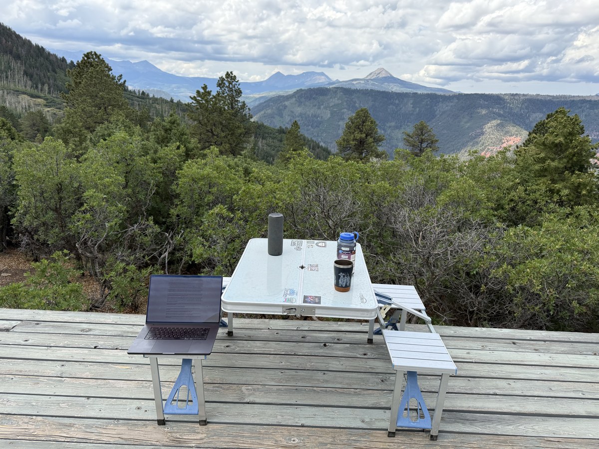A laptop and camp table set up on the yurt deck overlooking the San Juan Mountains, a speaker, water bottle, and coffee mug on the table