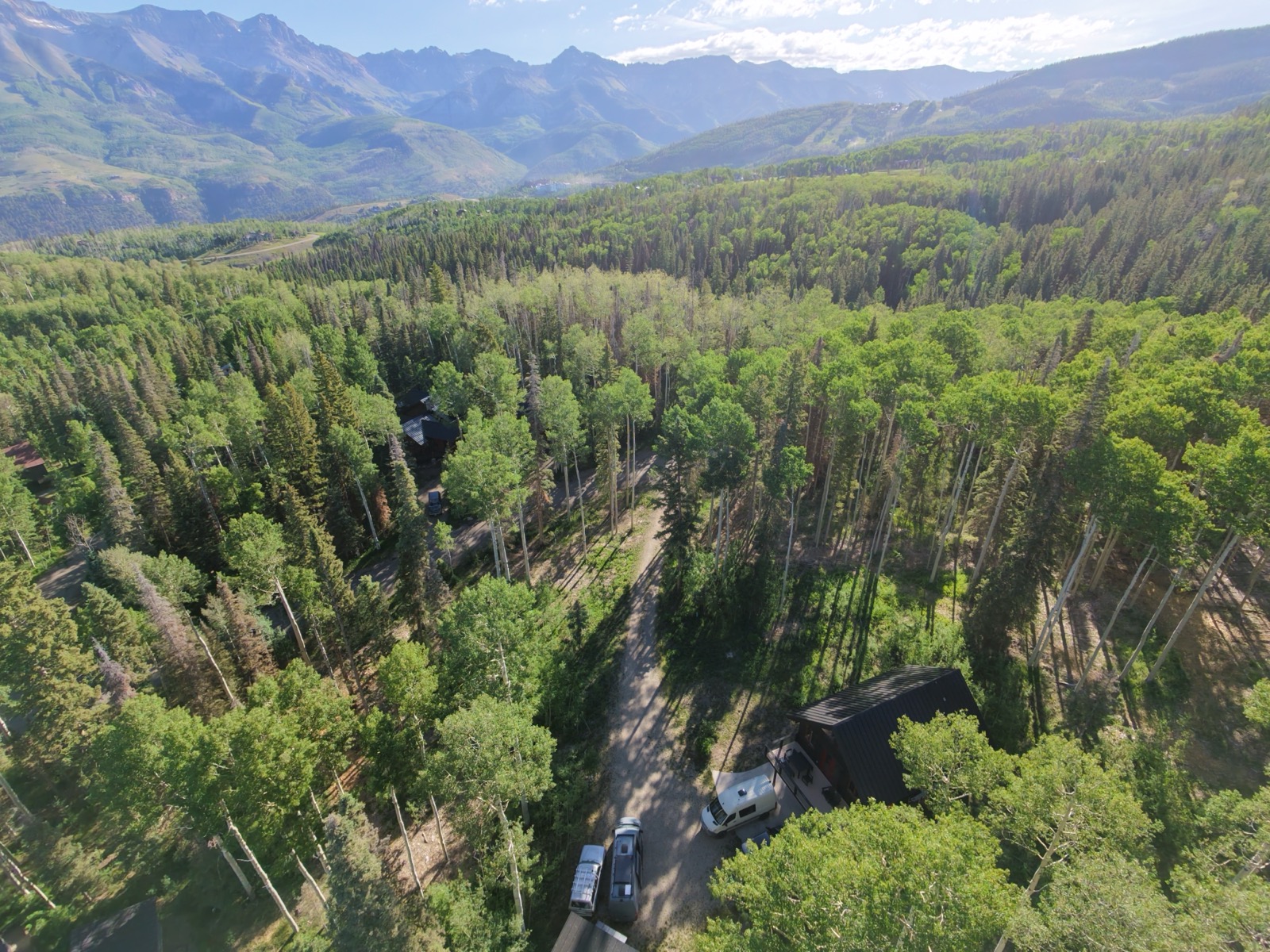 Aerial drone view of a dark-roofed cabin tucked into aspen and pine forest above Telluride, Colorado, with a Sprinter van parked in the driveway and the San Juan Mountains rising behind.