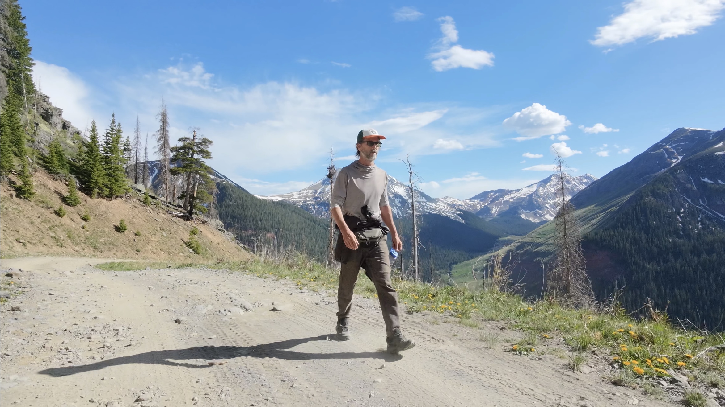 Layng walking a dirt road above a green valley, snow-capped San Juan Mountains rising on both sides under a blue sky.