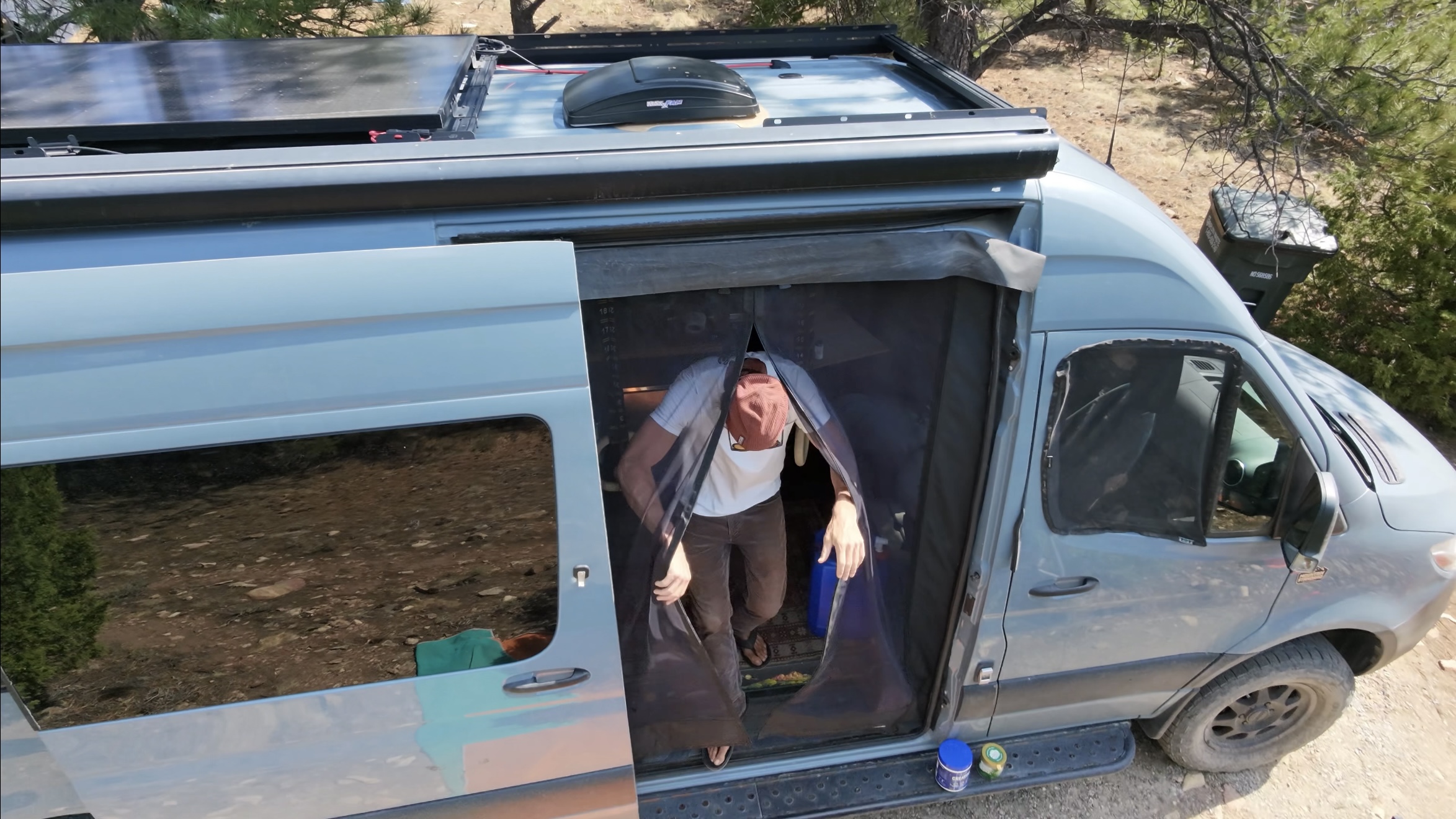 Aerial drone shot looking down at the Sprinter van, roof deck with solar panels and a rooftop box, standing through the open side door behind a mesh screen, holding a water jug