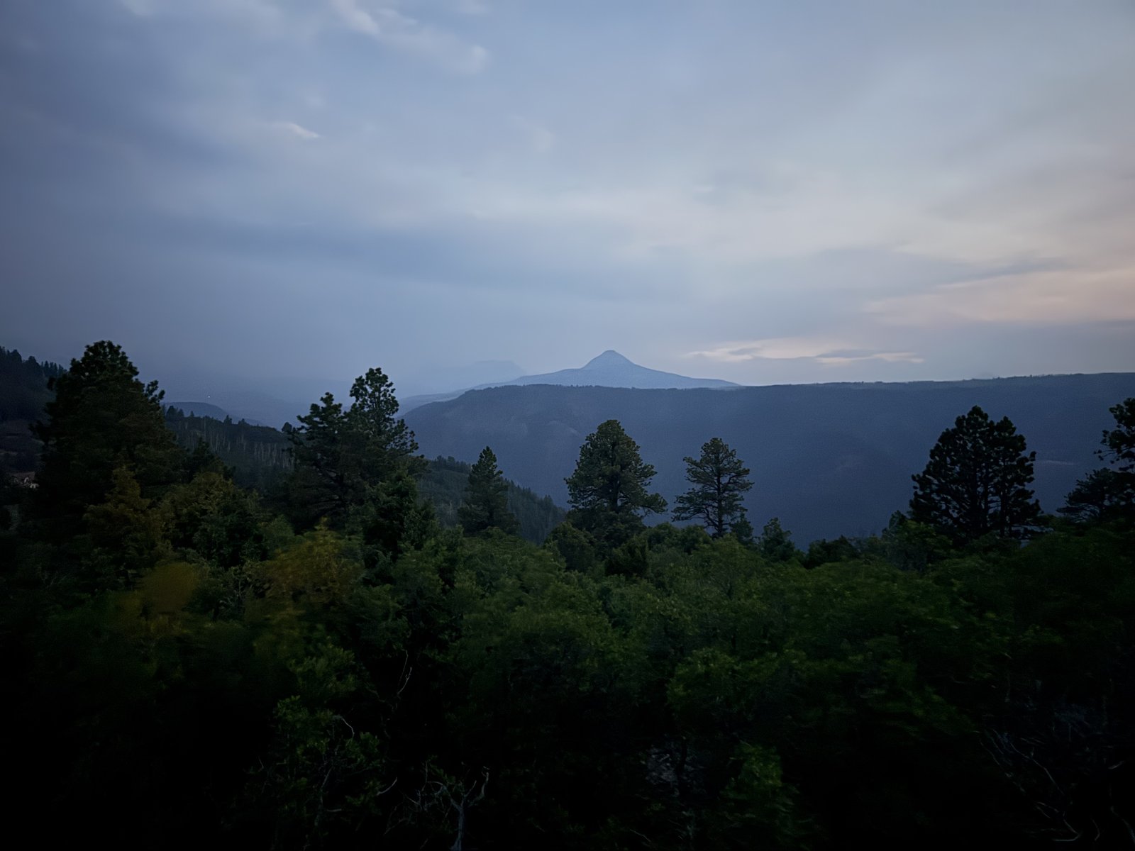 Dusk from the ridge above the yurt: dark pines in the foreground, hazy blue ridgelines beyond, and a lone conical peak silhouetted against the last light.
