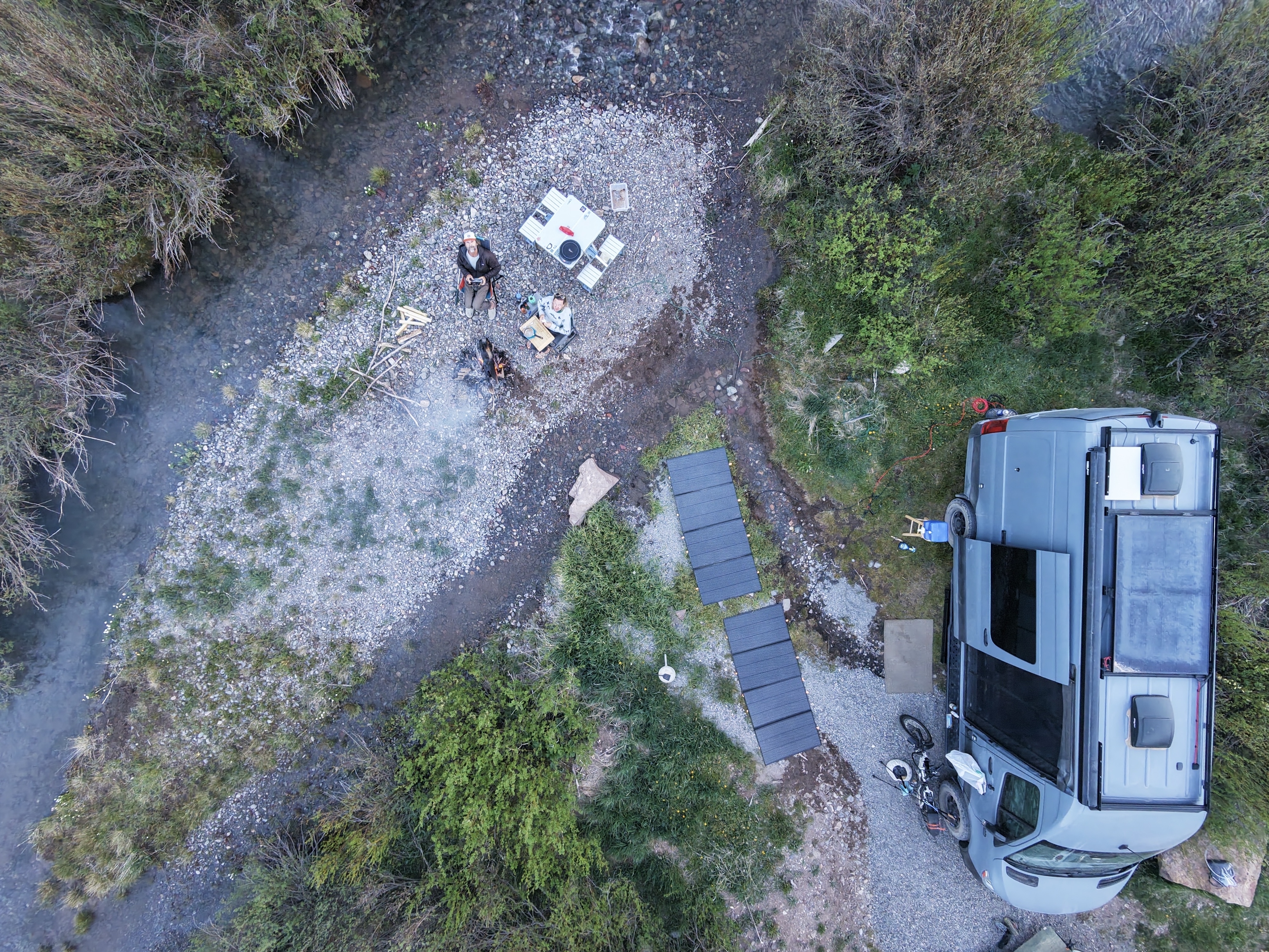 Aerial drone shot looking straight down at a campsite on a gravel bar beside a creek, two people by a fire, solar panels deployed, Sprinter van at right, San Juan Mountains, Colorado.