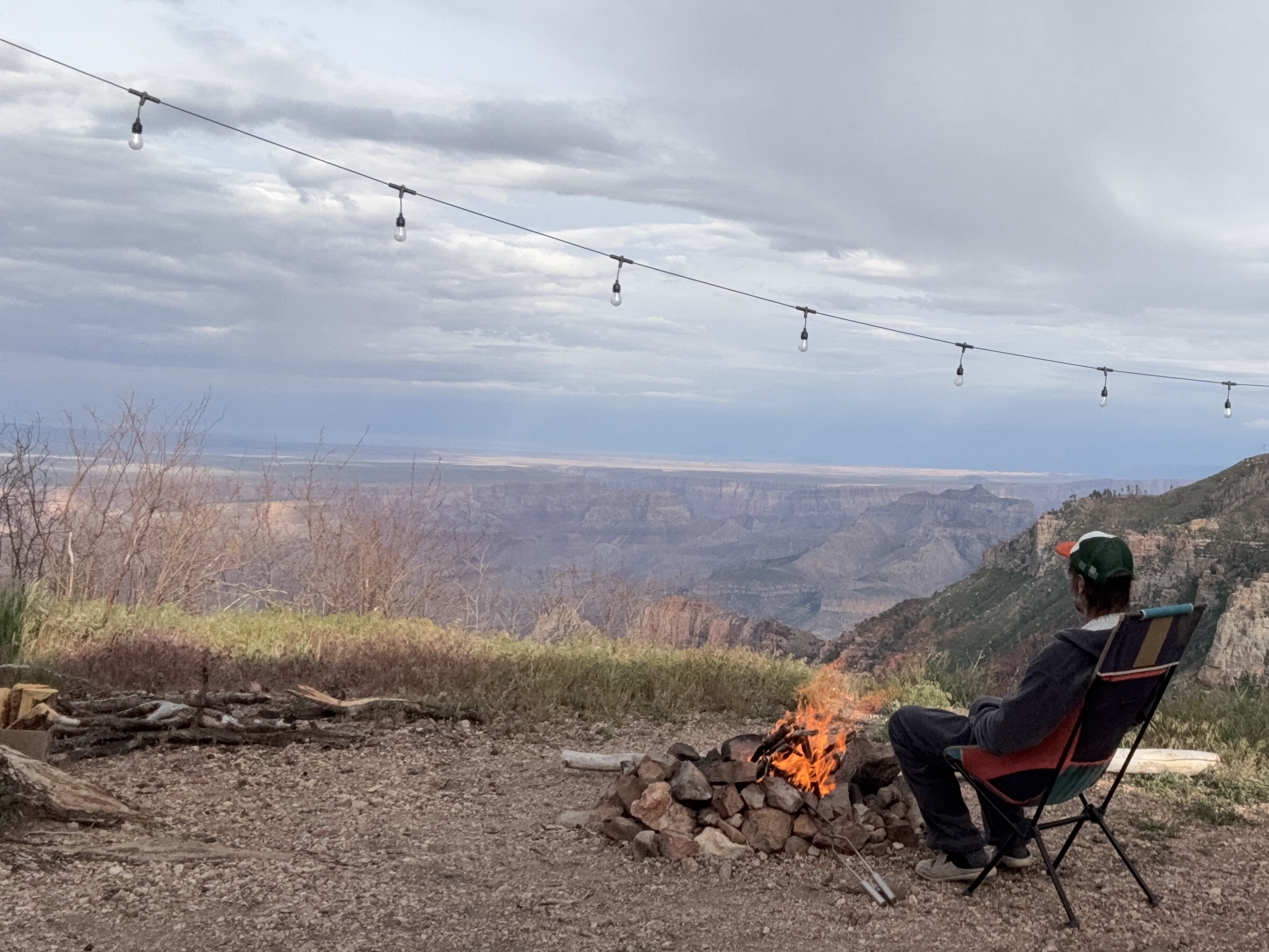 Sitting by a campfire at the edge of the Grand Canyon, string lights overhead, canyon stretching to the horizon