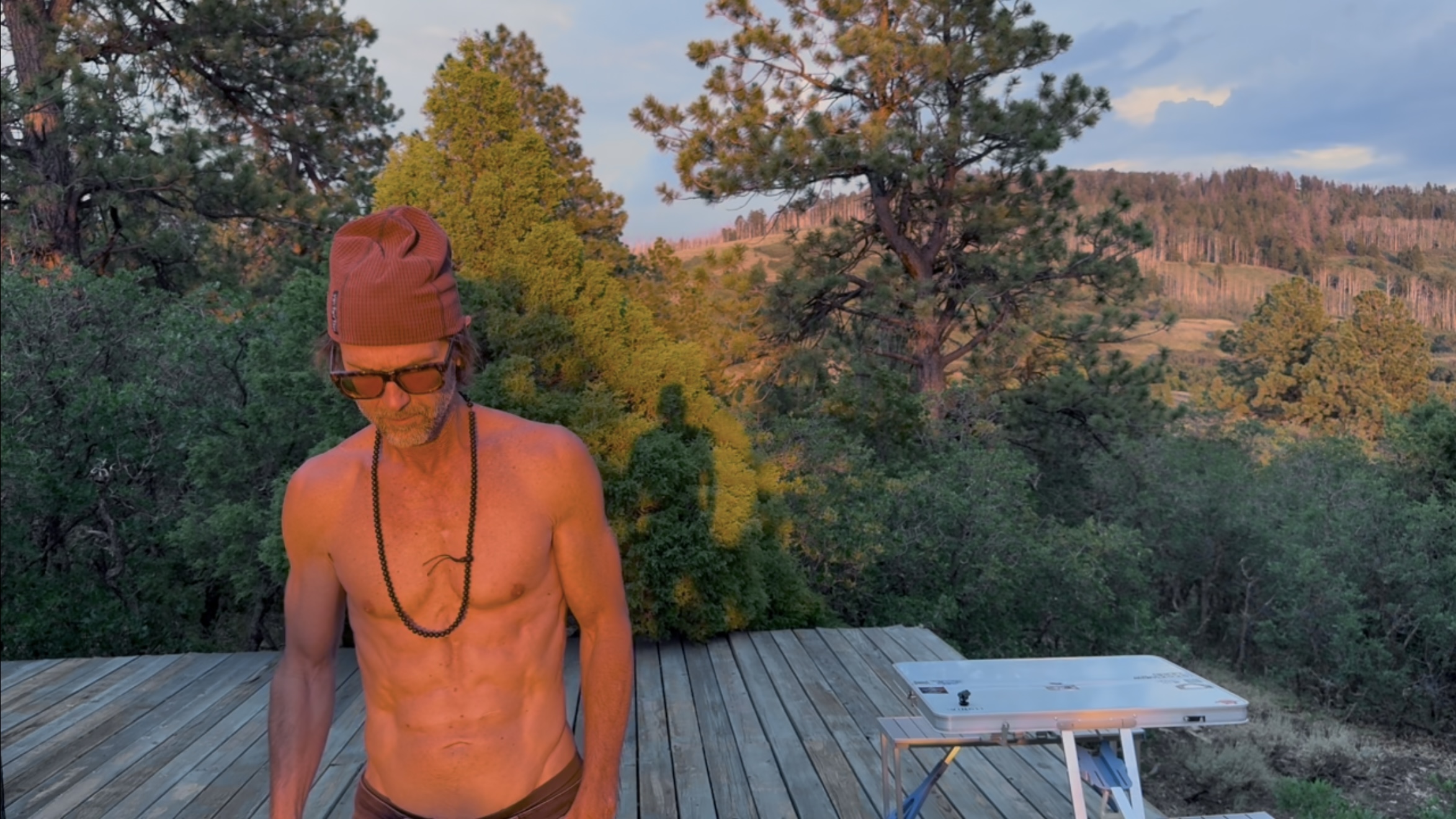 Portrait on a wooden deck at sunset, orange alpenglow lighting the pine forest and ridgeline behind, red beanie and sunglasses, a solar panel on a folding table nearby