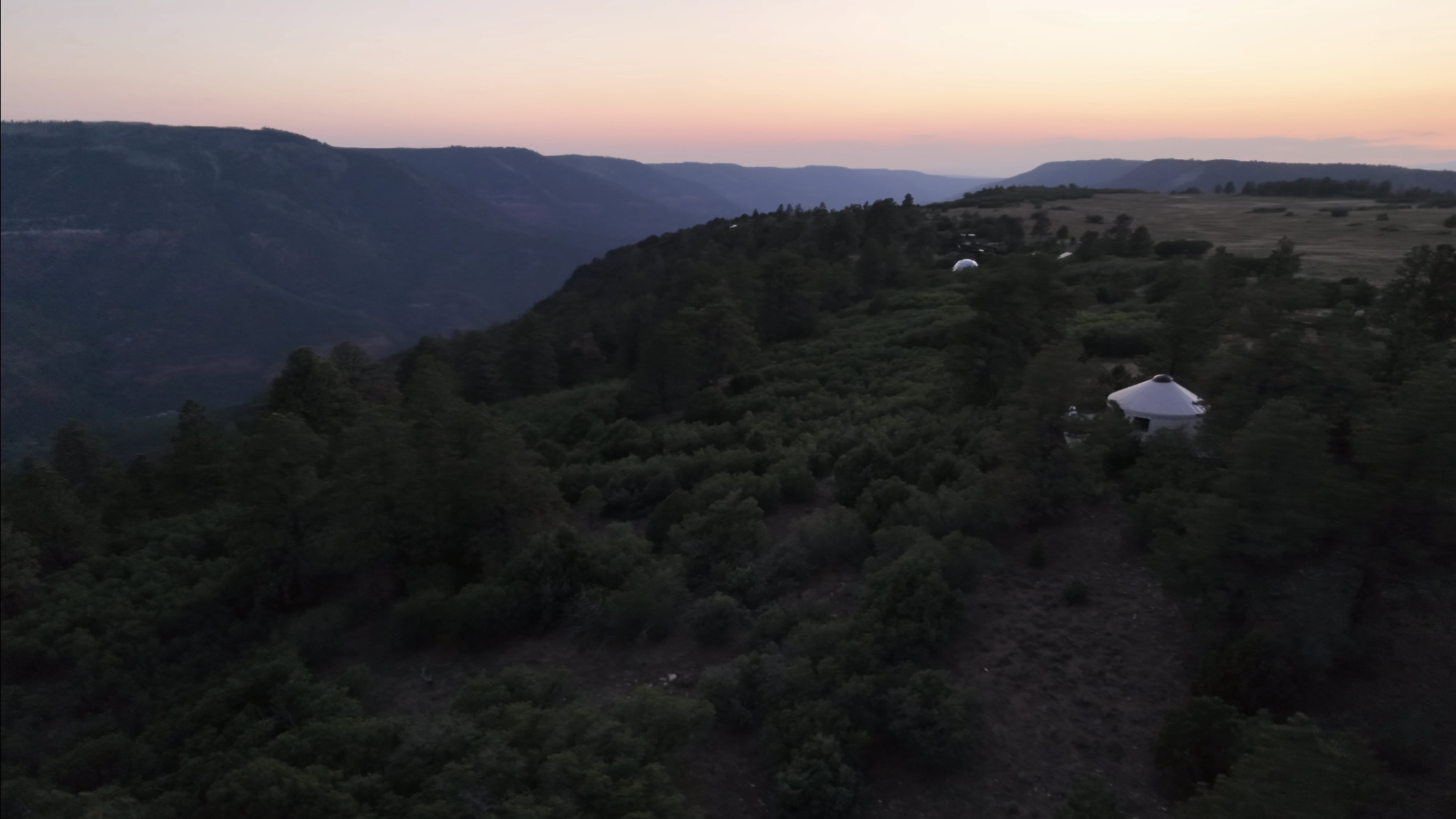 Aerial drone shot at dusk of a yurt and a dome-roofed structure sitting in a forest clearing on a ridge above a deep canyon, with a pink and orange sunset sky beyond distant mountains.