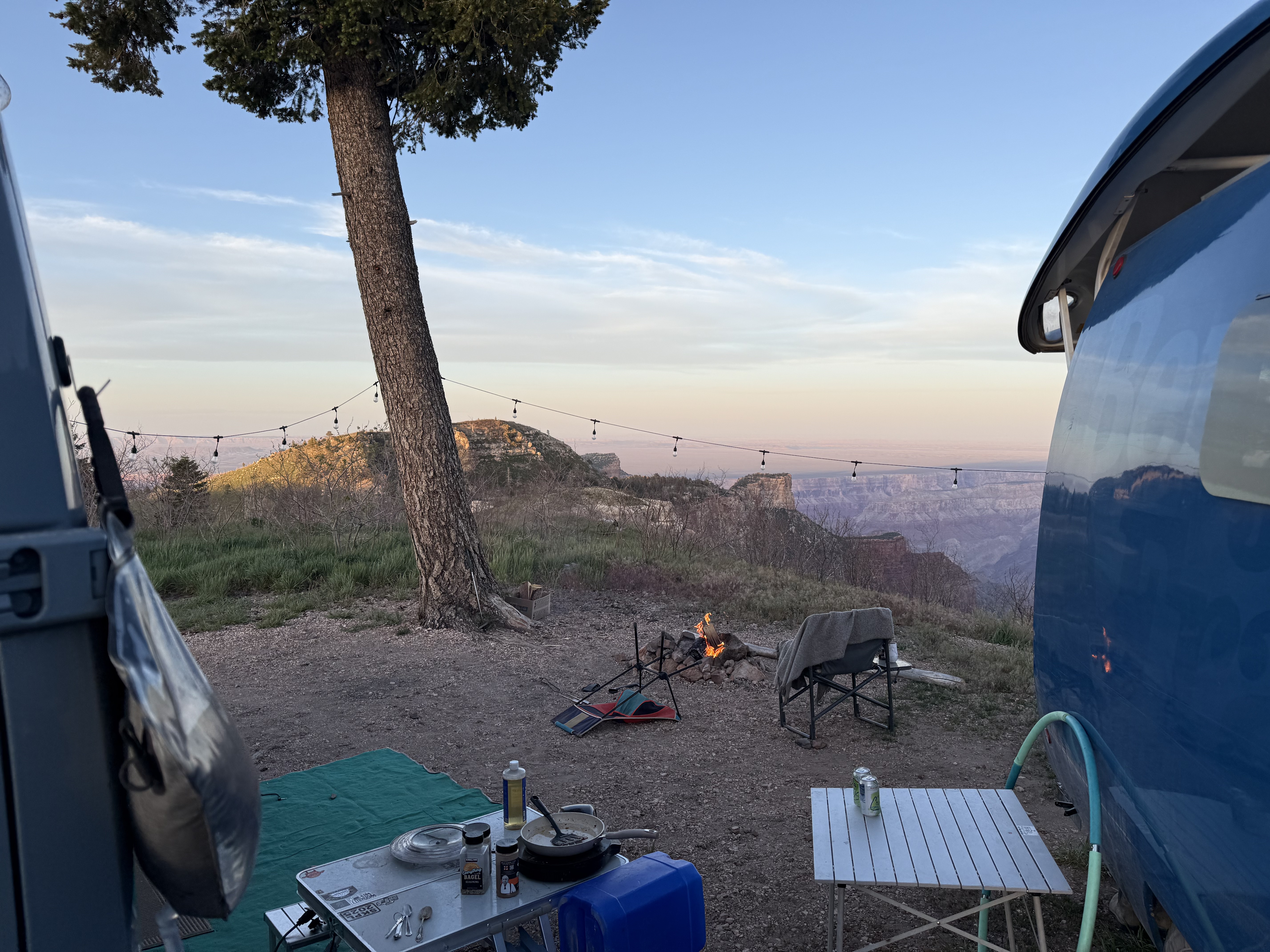 Last morning at the North Rim. Campfire between the van and an Airstream, string lights strung over the canyon at dusk.
