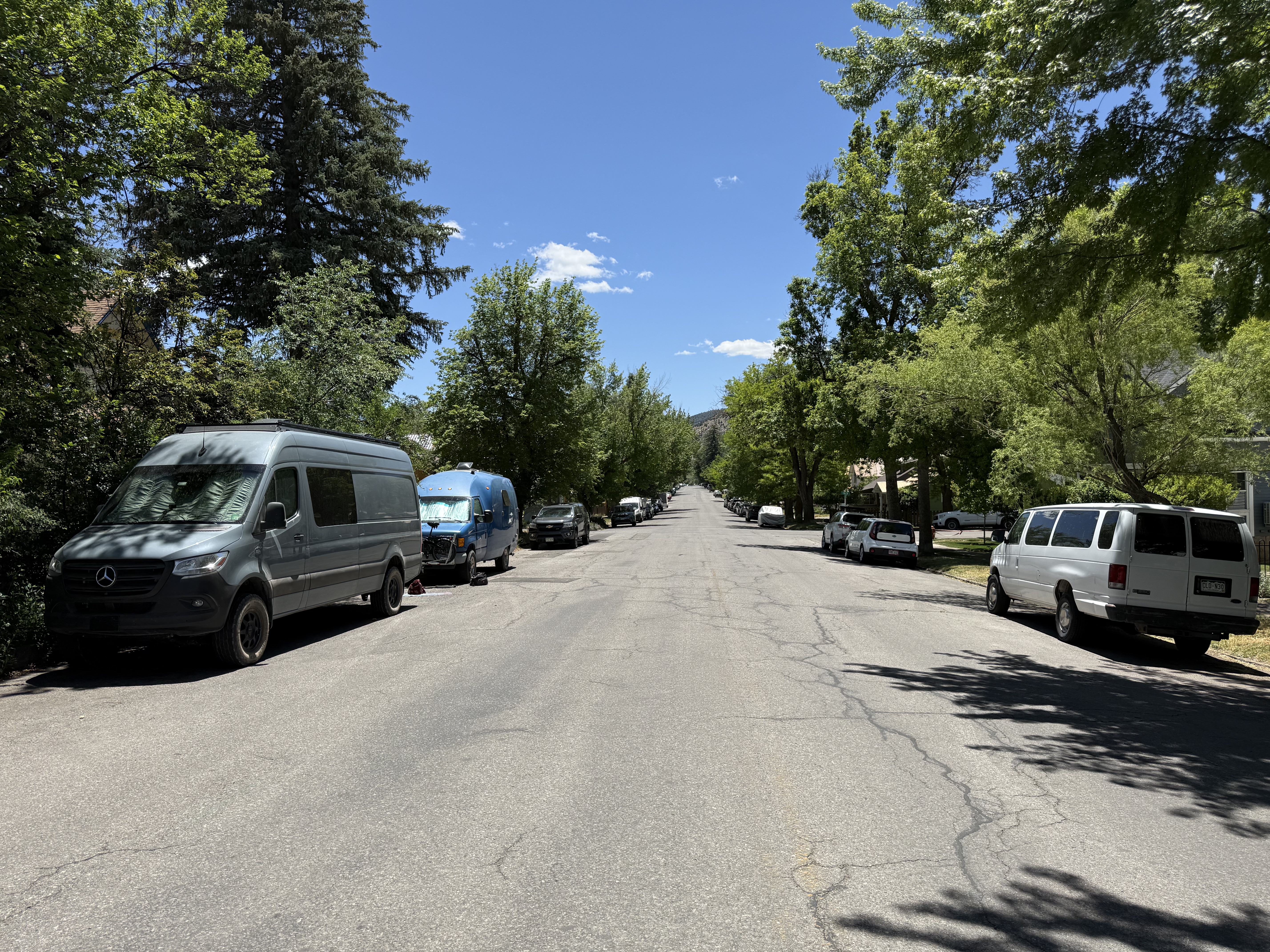 The Sprinter van parked on a tree-lined residential street in Durango, Colorado. Tall green trees shade both sides of the wide street under a bright blue sky, with mountains visible at the far end of the block.