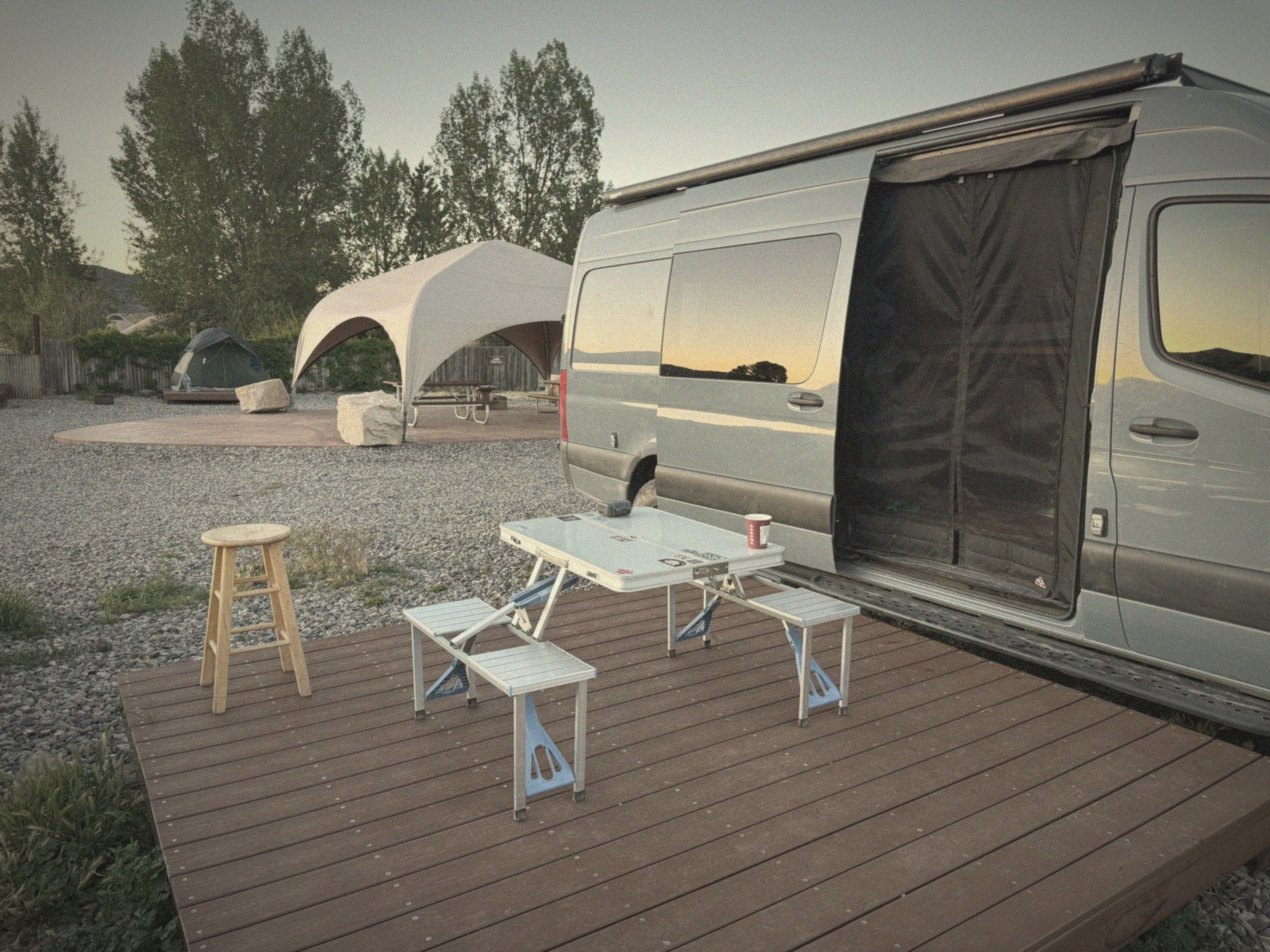 The Sprinter van parked beside a wooden tent platform at a Ridgway, Colorado campground, a portable table and stool set up on the deck at dusk.