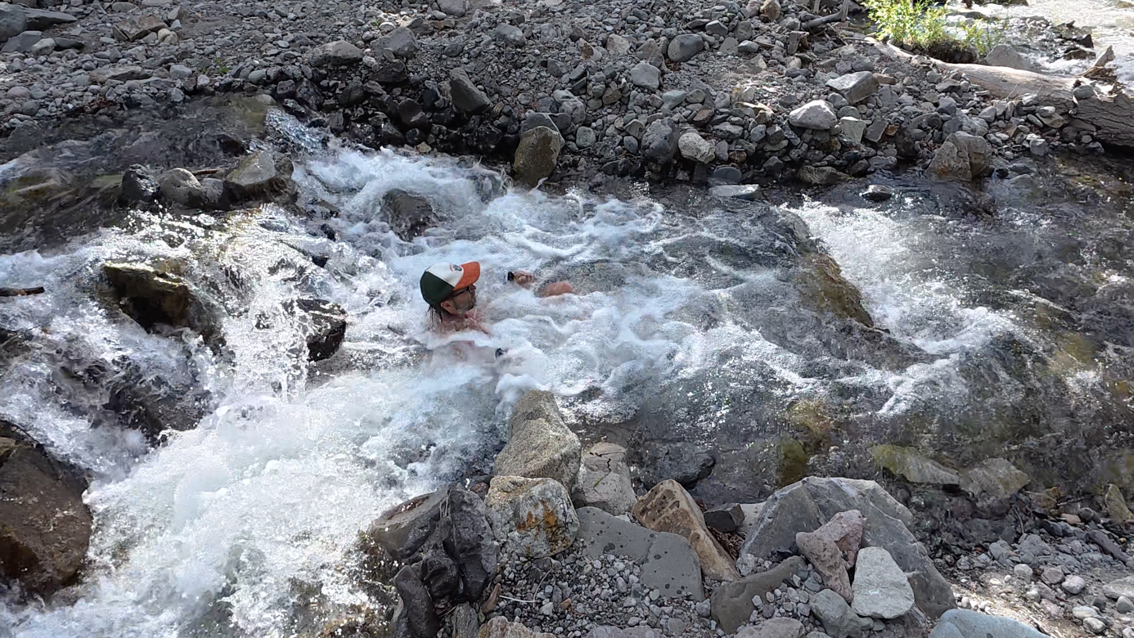 Layng submerged in a rushing rocky river, hat on, arms out, in a cold plunge in Ouray, Colorado.