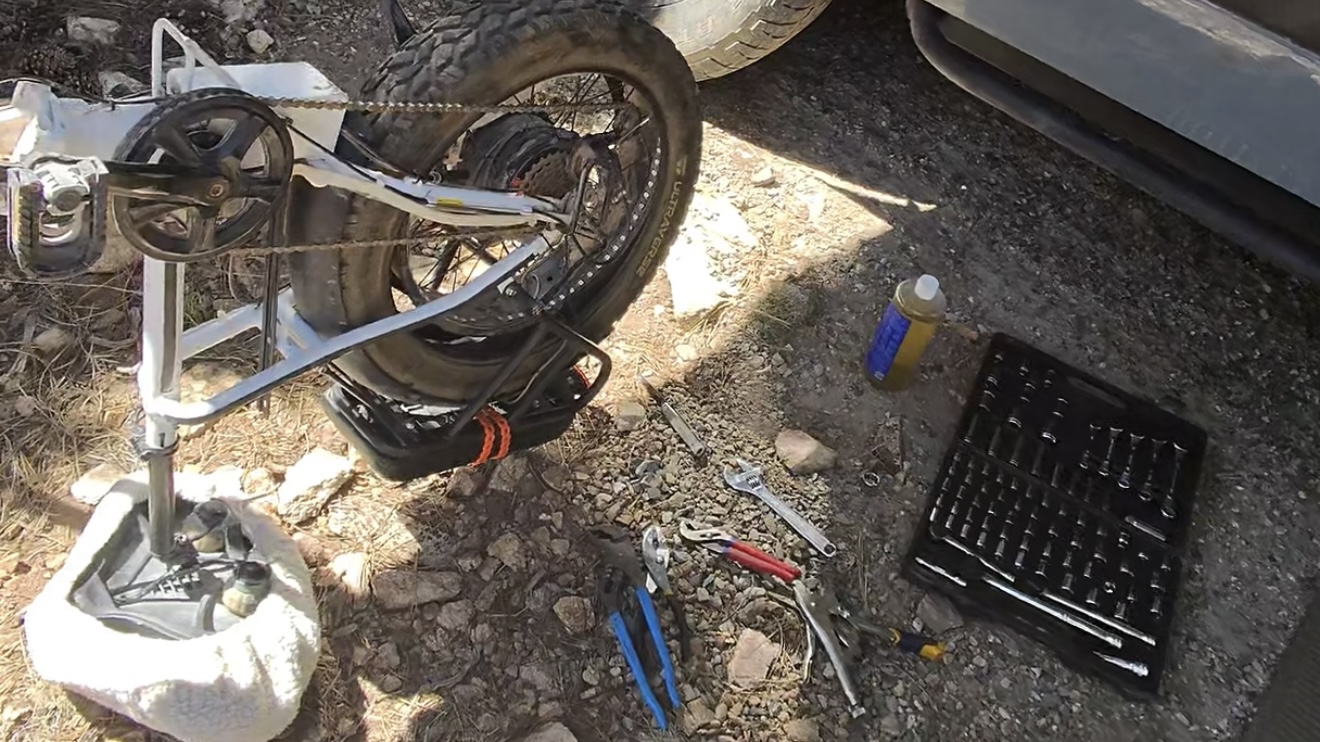 An e-bike up on a repair stand with its rear wheel exposed, a chain and sprocket visible, surrounded by wrenches, pliers, a socket set, and chain lube spread out on gravel.