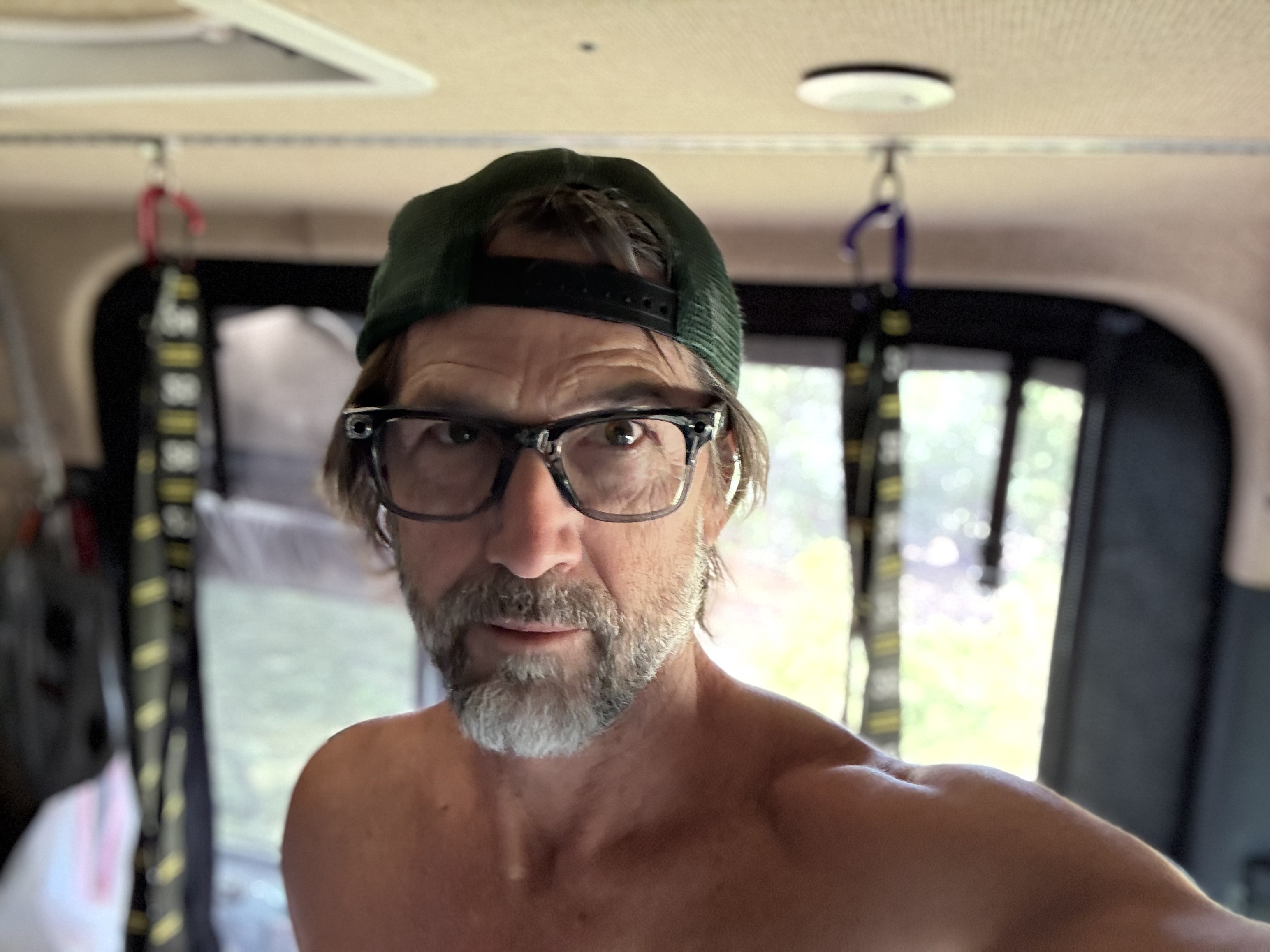 Selfie inside a van: a shirtless man wearing dark-framed Meta AI glasses and a backwards green trucker hat, looking directly at the camera.