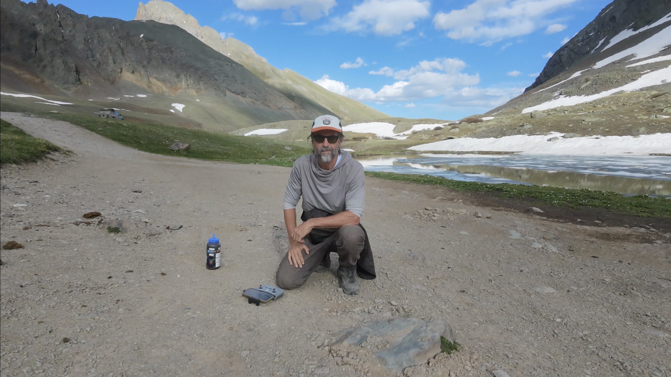 Layng kneeling at the edge of a half-frozen alpine lake at 12,000 feet, drone controller on the ground beside him, San Juan Mountains rising on all sides, Colorado.