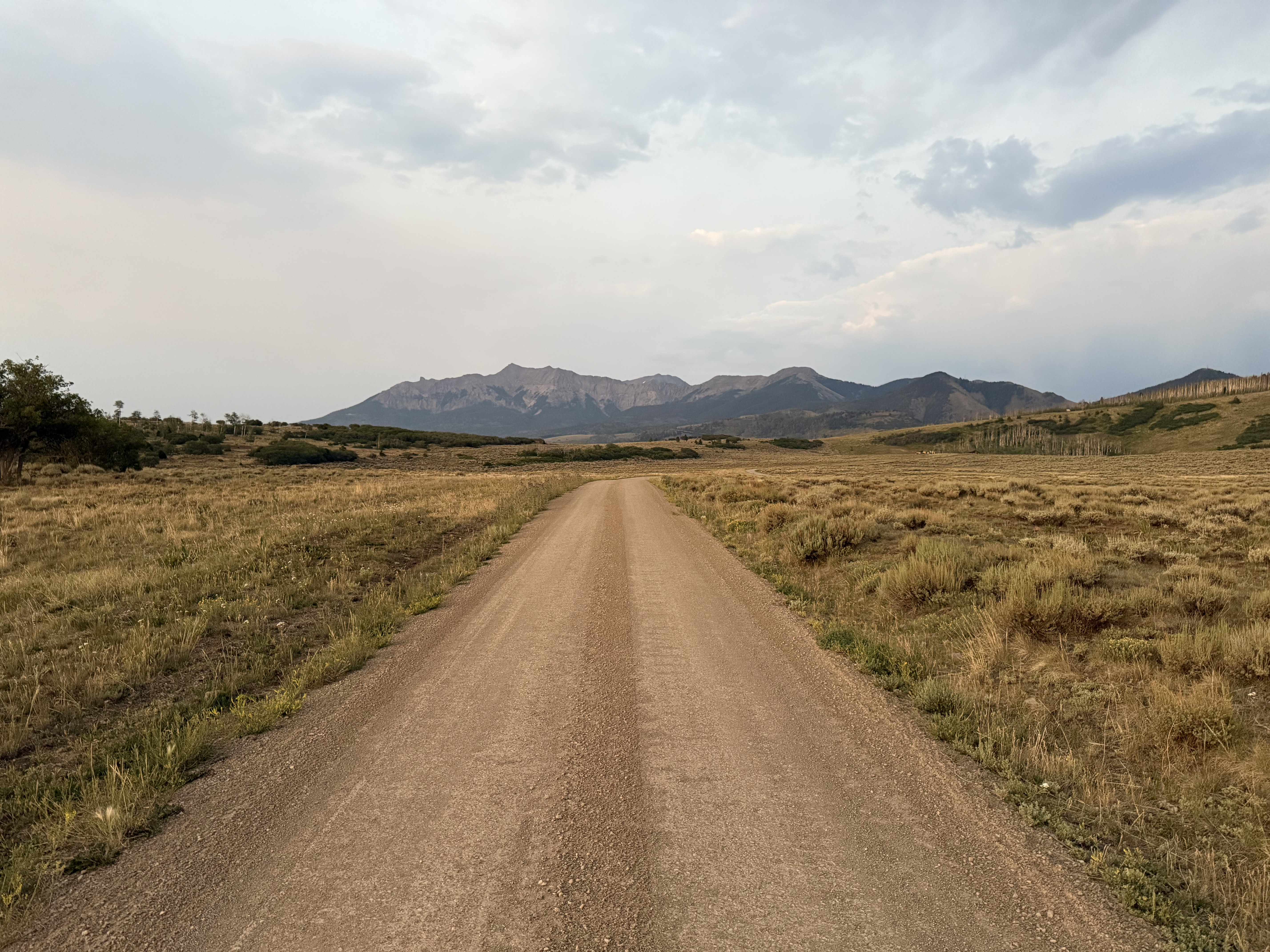 A straight dirt road stretching toward the San Juan Mountains at dusk, dry grass fields on either side