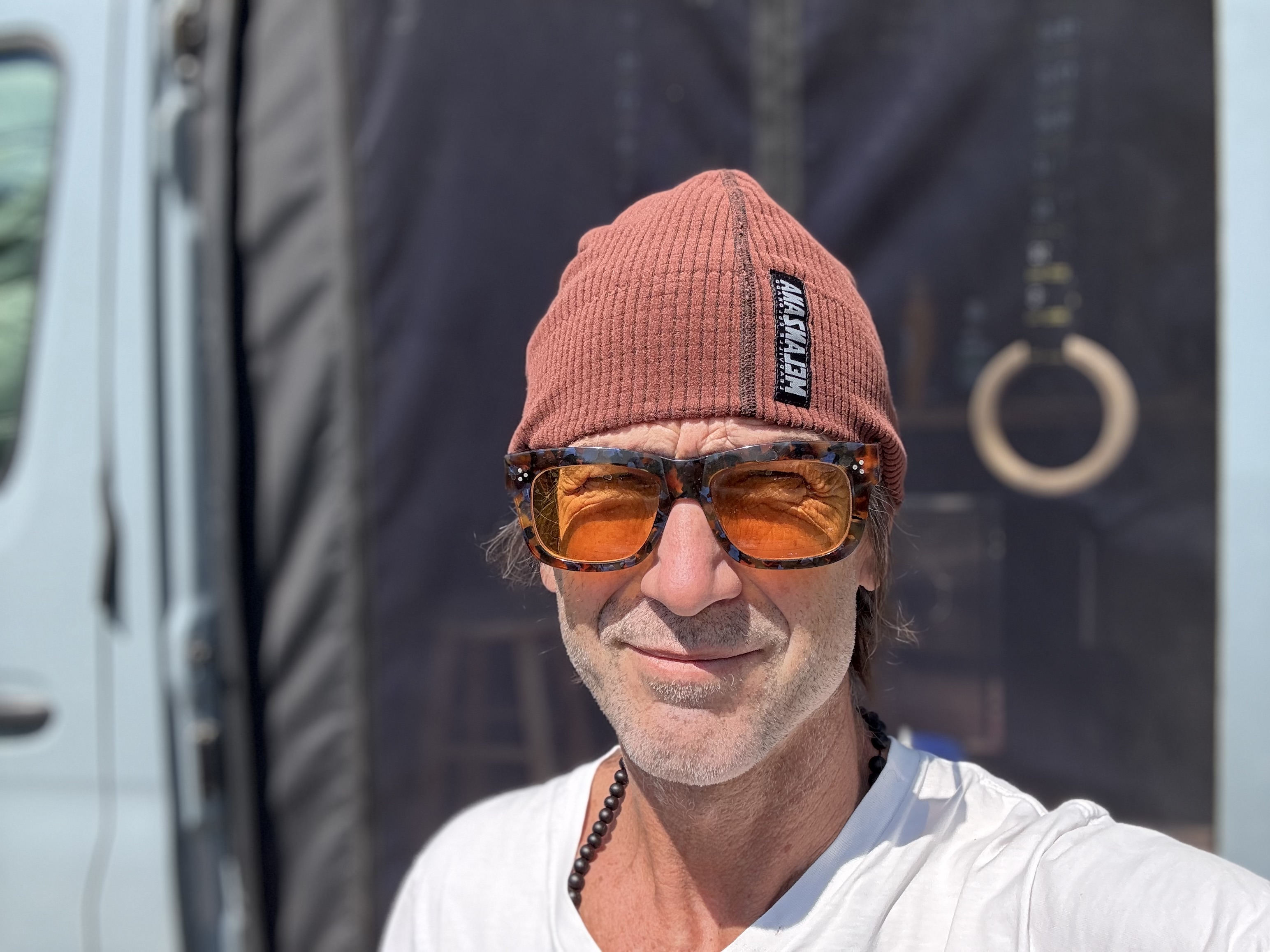 Selfie outside the van in Gunnison, Colorado: Melanzana beanie, orange-tinted sunglasses, short stubble after shaving the beard, gymnastic ring visible in the background.