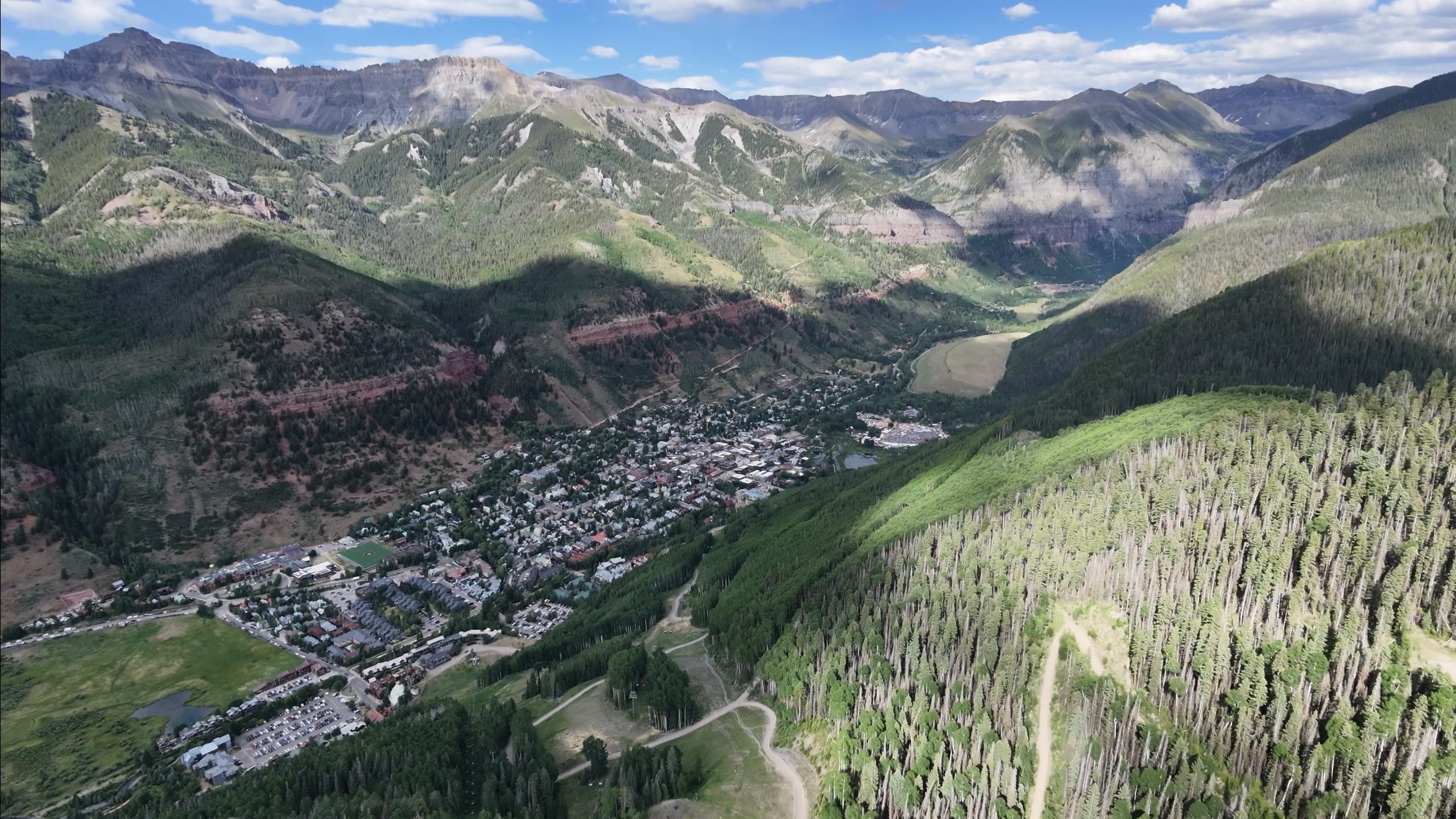 Aerial drone shot looking down a box canyon at the town of Telluride, Colorado, nestled between steep forested mountains with a red rock cliff band above town.