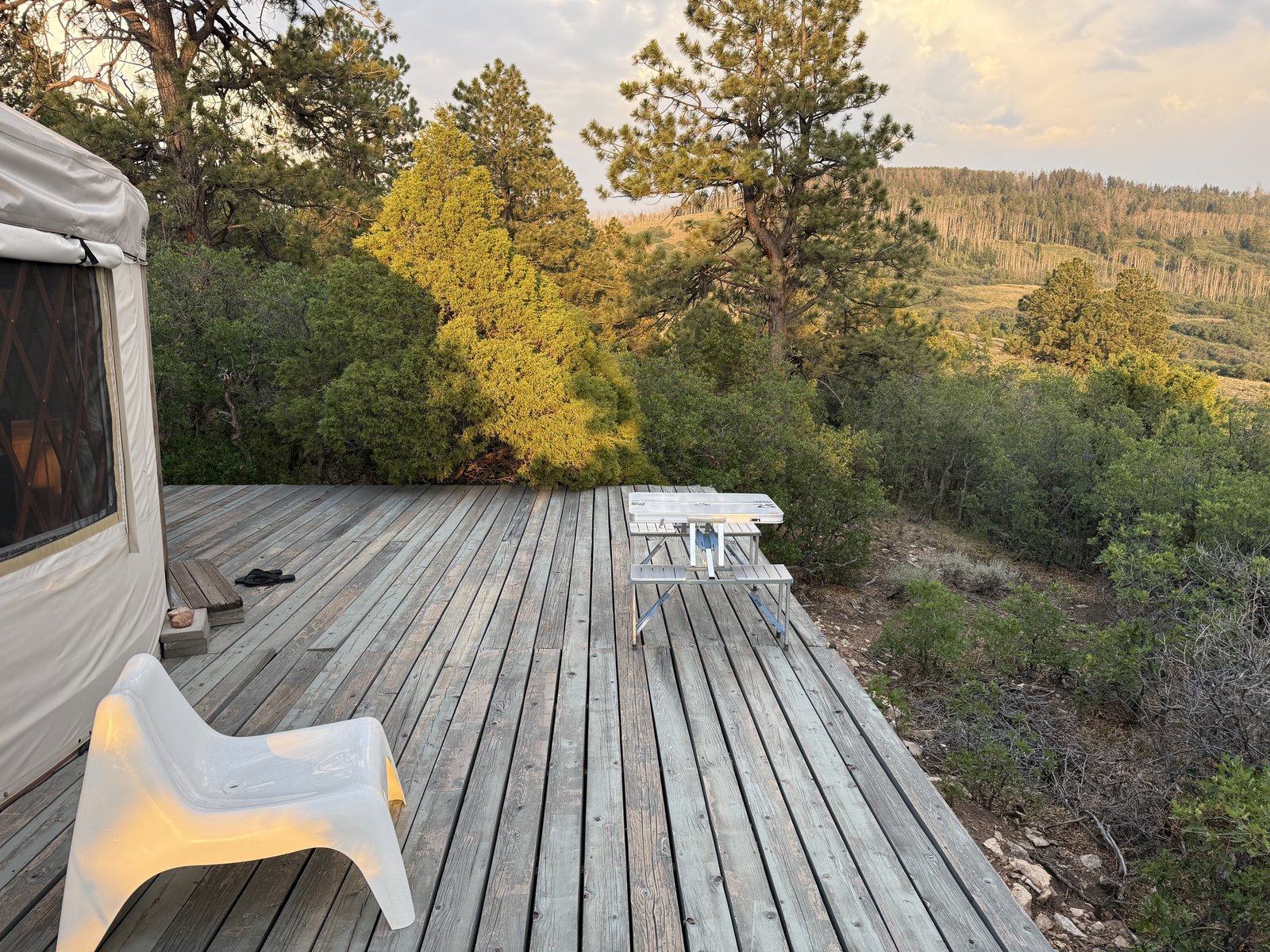 Golden evening light on the yurt deck: weathered gray boards, a white chair, the folding picnic table, and sunlit pines dropping away toward the valley.
