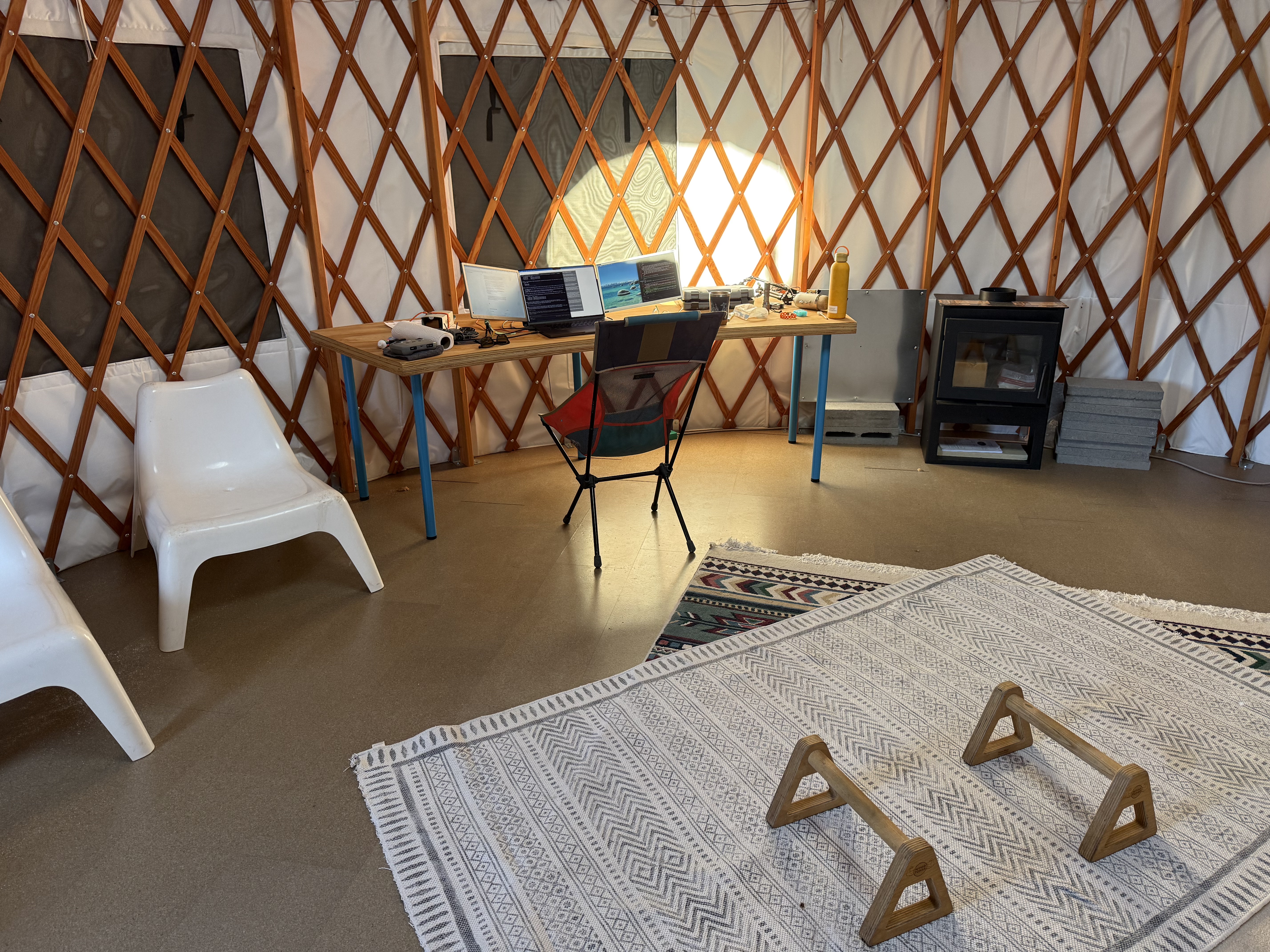 Interior of a yurt in Telluride, Colorado: a work desk with two laptops and a glowing lamp against the lattice walls, a wood stove to the right, a patterned rug on the floor with two wooden paralettes, and two white chairs to the left.
