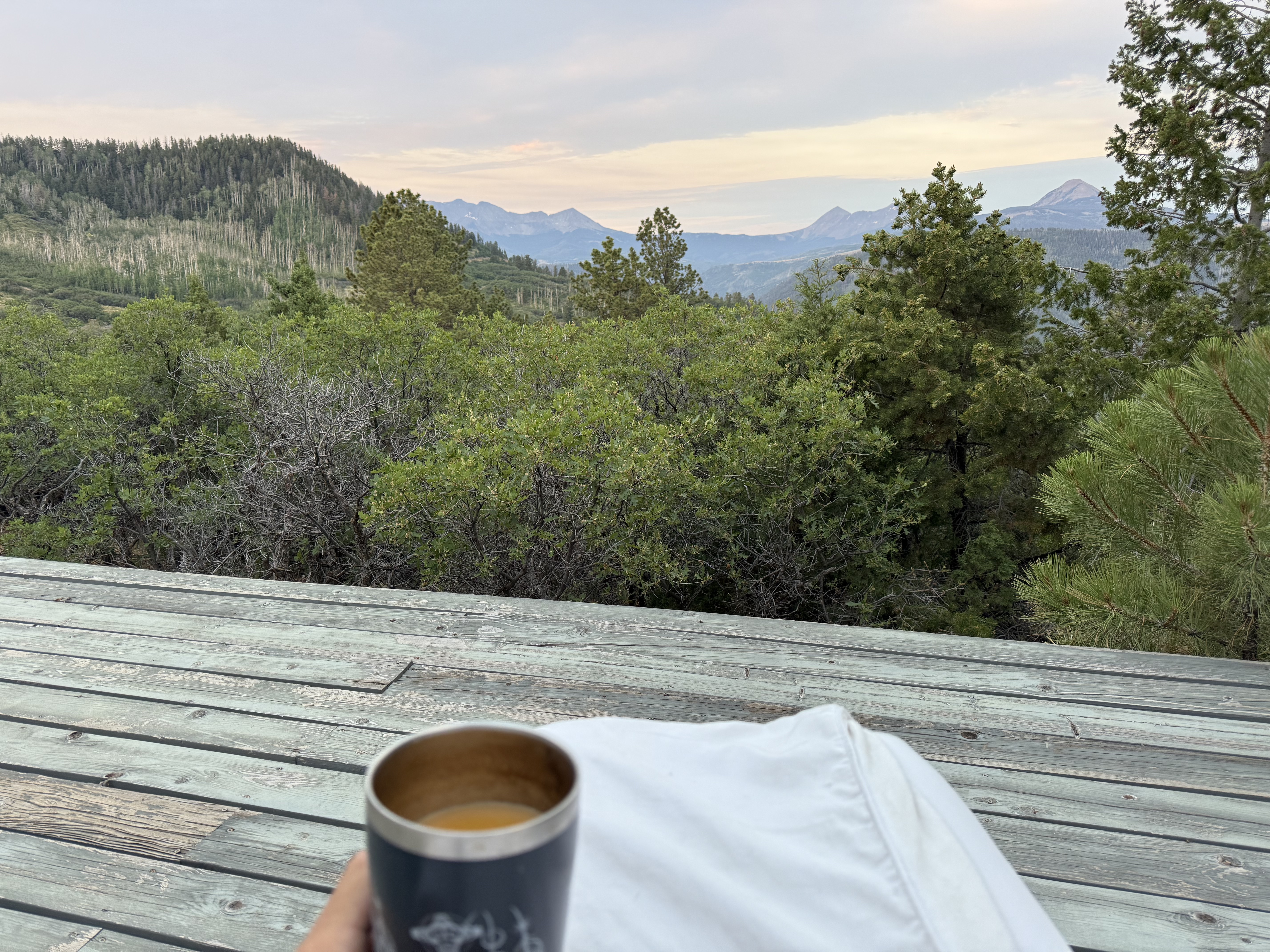A mug of coffee with ghee held up against a dawn view of the San Juan Mountains from the deck, a white bathrobe sleeve in frame