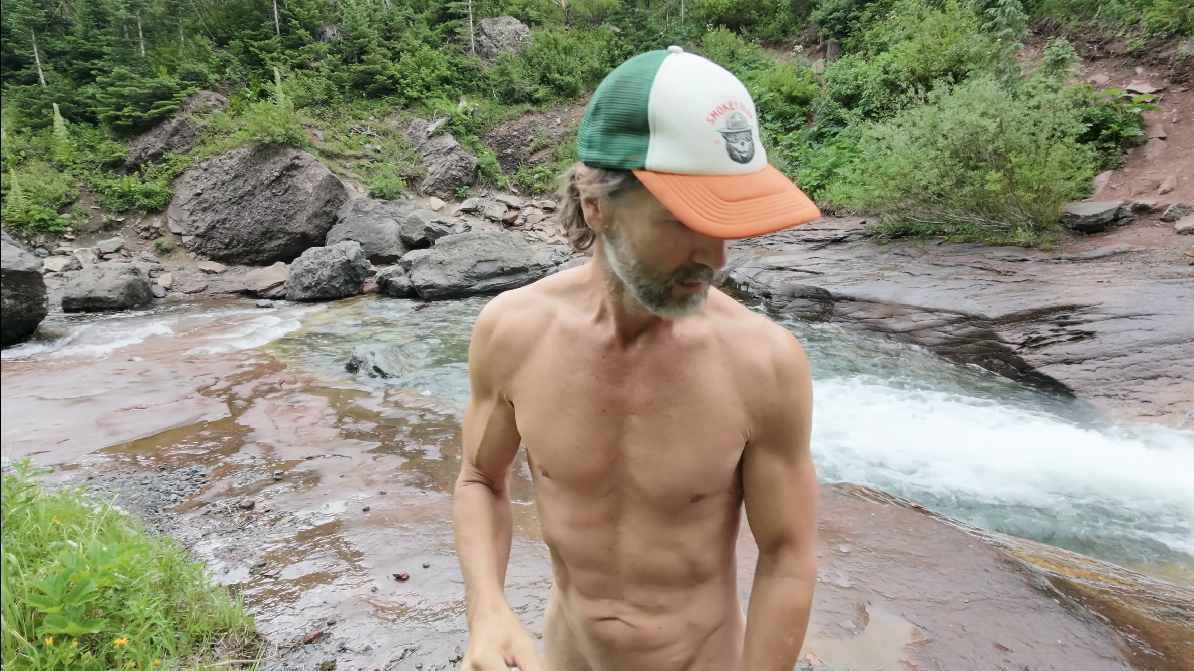 Layng exiting a cold plunge in an alpine river near Telluride, Colorado, wearing a Smokey Bear trucker hat, rushing water and boulder-strewn banks behind him.
