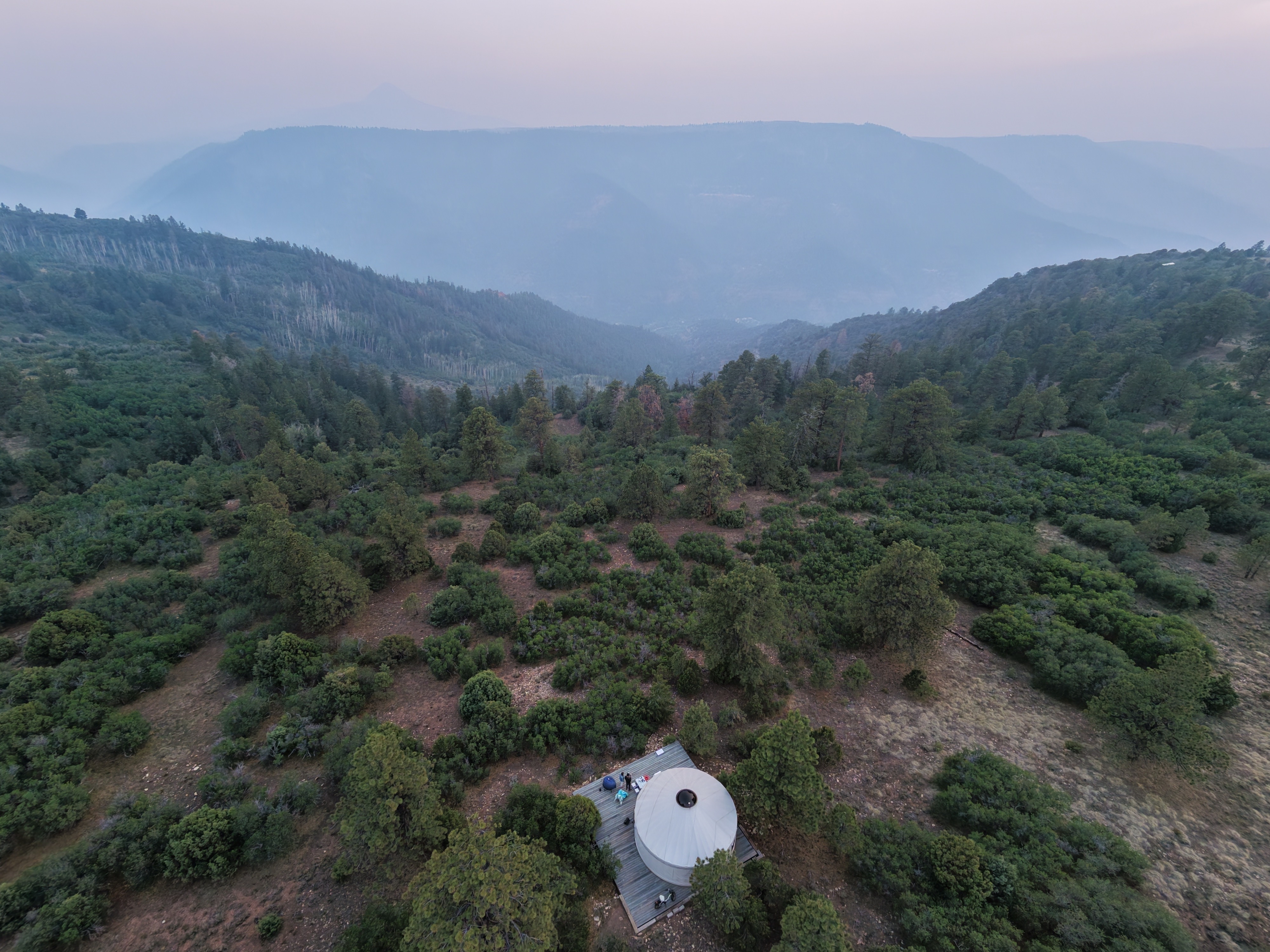 Aerial drone shot at dusk of a dome-roofed yurt on a deck in a forest clearing near Telluride, Colorado, with distant mountains hazed over by wildfire smoke drifting in from Utah.