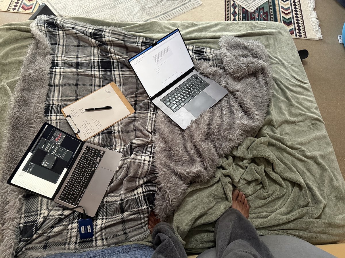 Two MacBooks and a clipboard of handwritten notes spread across blankets in the yurt, seen from above, bare feet at the edge of the frame.