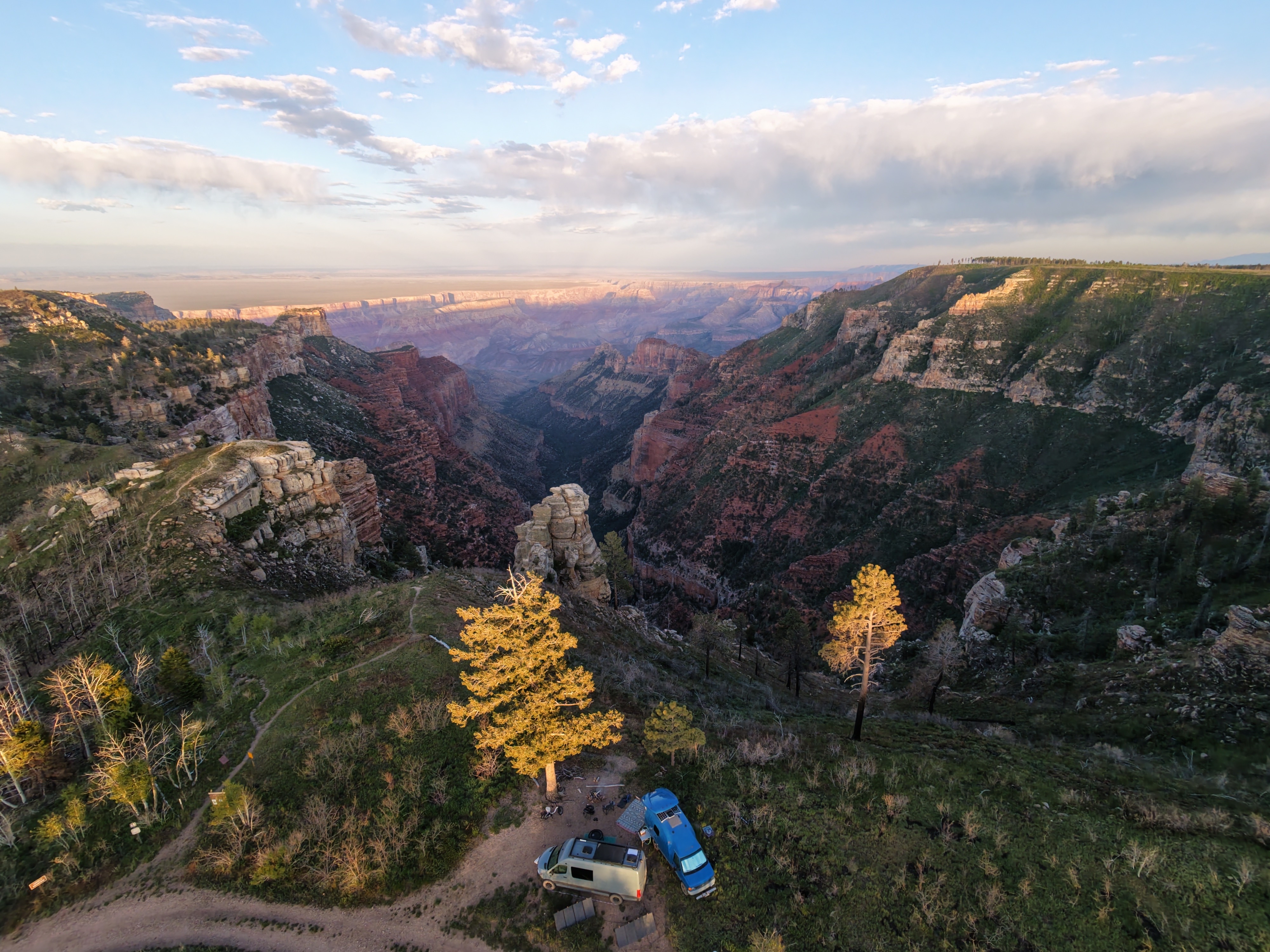 Aerial drone view of two vans camped on the rim of the Grand Canyon at golden hour, ponderosa pines lit gold