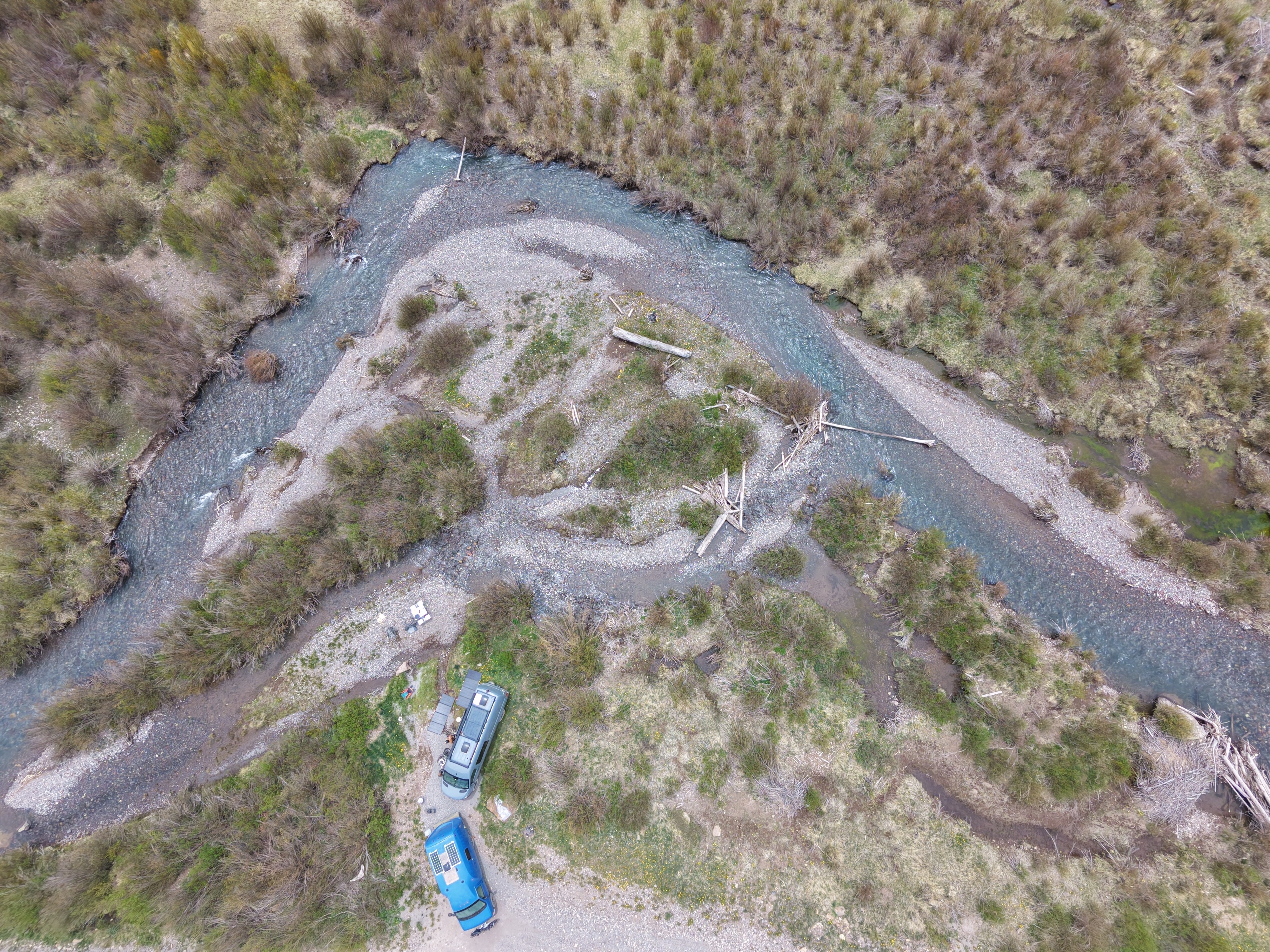 Aerial drone shot looking down at two vans camped on a gravel bar where a blue-green snowmelt creek bends through the San Juan Mountains, Colorado.